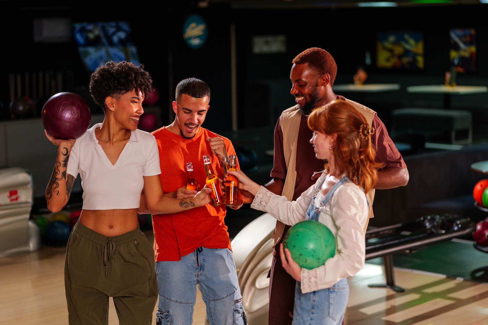 Four young adult friends are toasting and holding bowling balls in a bowling alley, enjoying their leisure time together